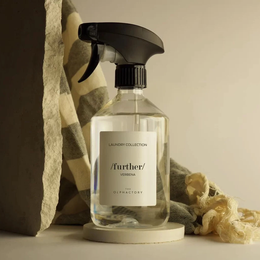 Bruma textil Verbena Further en botella con rociador, junto a textiles y sobre una base redonda