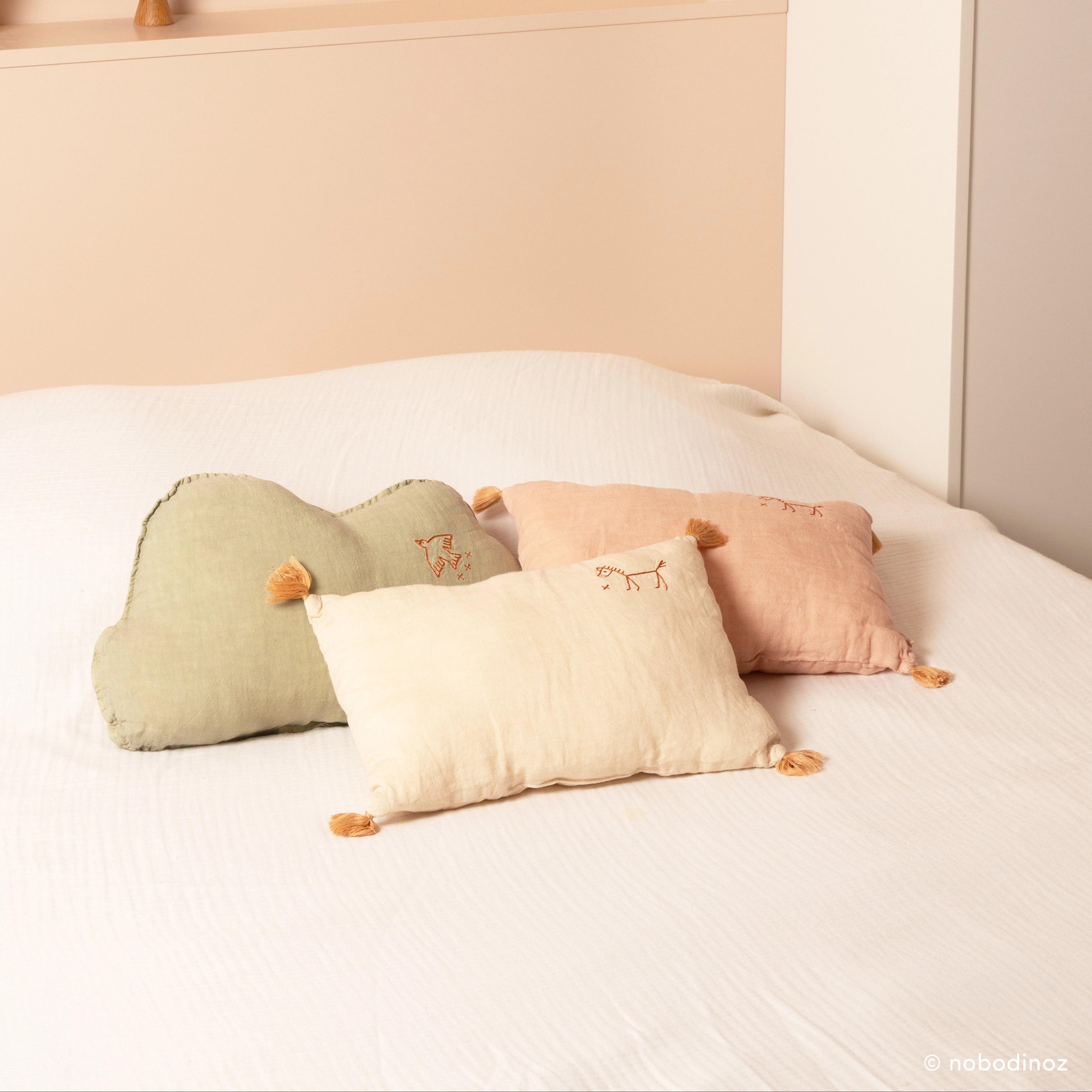 Cojines rectangulares con borlas y bordados sobre una cama; el cojín claro está en primer plano.