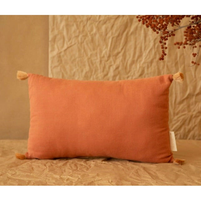 Cojín Toffee rectangular con pompones en las cuatro puntas, visto de frente sobre una cama.